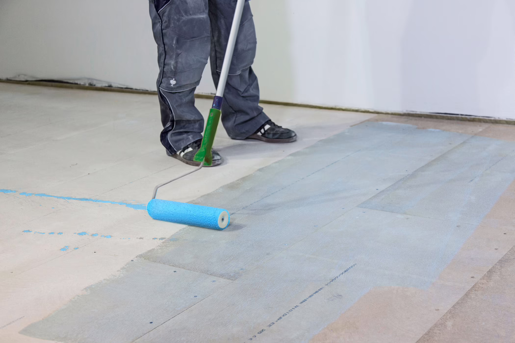 Die Unterschenkel und Füße eines Handwerkers sind zu sehen, während er mit einer langstieligen Farbrolle einen hellblauen Grundanstrich auf einem Holzunterboden aufträgt. Die Person trägt dunkle Arbeitshosen und schwarze Turnschuhe; die Rolle hinterlässt einen feuchten blauen Film auf den Dielen in einem Innenraum mit weißen Wänden. Das Bild konzentriert sich auf die Bodenvorbereitung, Werkzeuge und Farbspritzer.