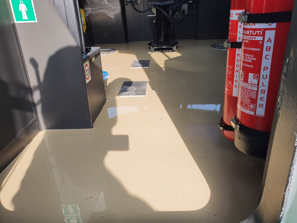 Der Boden eines Raums ist mit Wasser bedeckt; an der rechten Wand sind Feuerlöscher angebracht, außerdem sind Sicherheitsschilder und Maschinen zu sehen. Auf der nassen, beigen Bodenfläche spiegeln sich Sonnenlicht und Schatten.