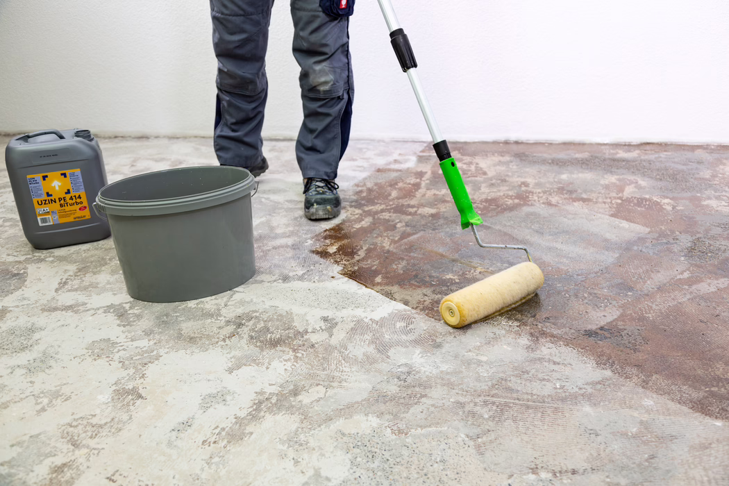 UZIN_Application Image_PE 414_9406_2018-11 Die unteren Beine und Füße eines Handwerkers sind zu sehen, während er mit einer langstieligen Farbrolle eine braune Grundierung beziehungsweise Klebeschicht auf einen nackten Betonboden aufträgt. Ein grauer Kunststoffeimer und ein grauer Kanister mit der Aufschrift "UZIN PE 414 Biturbo" stehen in der Nähe an einer weißen Wand. Der Handwerker trägt graue Arbeitshosen und schwarze Sicherheitsschuhe; der Boden zeigt verbliebene Kleberreste und geschliffenen Beton, während die Rolle die Flüssigkeit gleichmäßig verteilt, um den Untergrund vorzubereiten.
