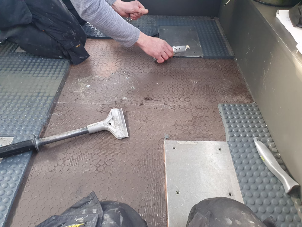 Zwei Personen sind damit beschäftigt, braune sechseckige Bodenfliesen mit Metallschabern zu entfernen. Einige Fliesen wurden bereits abgenommen, und auf dem Boden liegen verschiedene Werkzeuge verstreut.