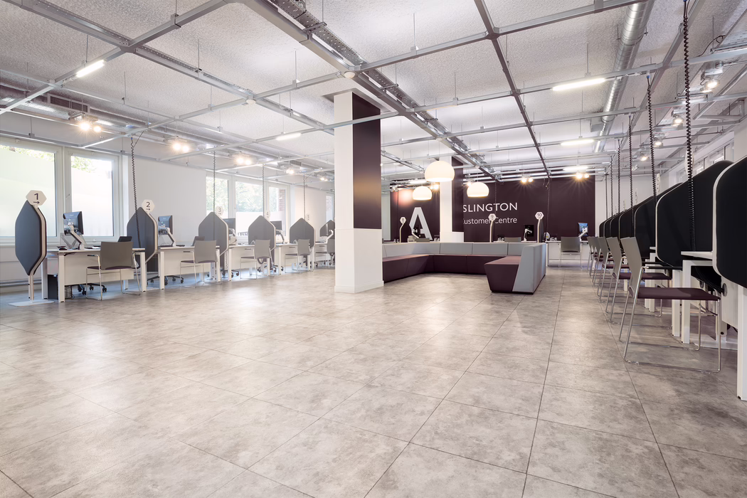 Ein heller, moderner Kundencenter-Saal mit weitläufigem, offenem Grundriss und hellgrauem Fliesenboden. Links und rechts reihen sich weiße Service-Schalter mit hohen, dunklen Sichtschutzwänden und nummerierten Schildern über jedem Arbeitsplatz; an jedem Schalter steht ein Monitor und ein einfacher Besucherstuhl. In der Raummitte befindet sich eine große, quadratische Stütze sowie eine modulare Sitzlandschaft in Lila- und Grautönen, über der hängende Pendelleuchten angebracht sind. Die Decke zeigt ein offenes Träger- und Kabelraster mit Beleuchtungsschienen und sichtbaren Lüftungsrohren, was dem Raum ein zeitgemäßes, leicht industrielles Erscheinungsbild verleiht. Große Fenster an der linken Seite lassen natürliches Tageslicht einfallen; der Bereich wirkt sauber, minimalistisch und derzeit unbesetzt.