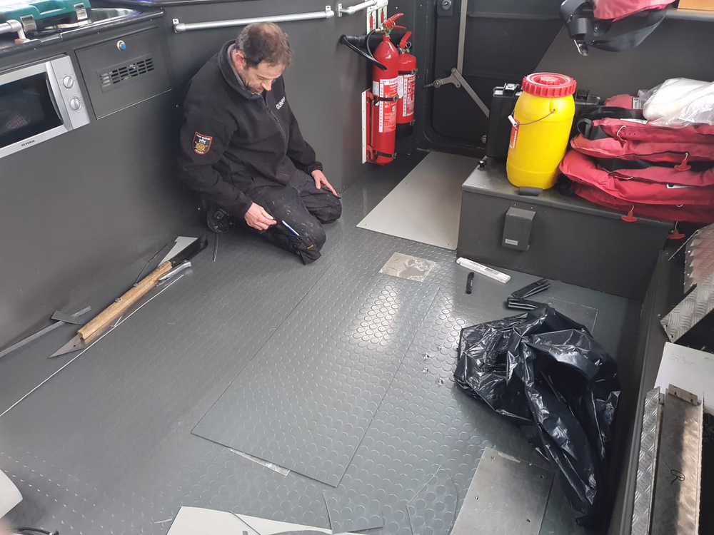 Eine Person kniet auf dem grauen, gemusterten Boden eines Raums, in dem Feuerlöscher, ein gelber Behälter, Werkzeuge und Material verstreut liegen, und scheint Reparatur- oder Installationsarbeiten durchzuführen.