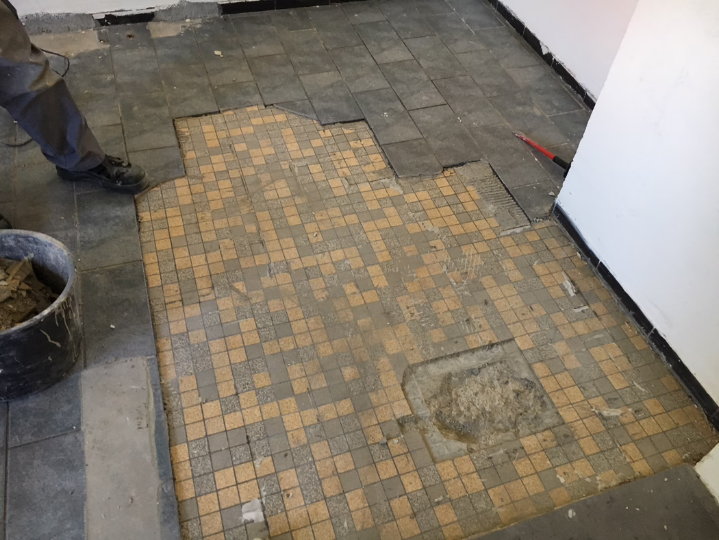 Teilweise entfernter Fliesenboden in einem Raum während Renovierungsarbeiten: Im Vordergrund sind kleine beige‑graue Mosaikfliesen mit abgenutzten und fehlenden Stellen zu sehen; größere dunkle rechteckige Randfliesen wurden gelöst und geben Kleber und Unterboden frei. Links sind die Beine und schwarzen Arbeitsschuhe eines Handwerkers neben einem Eimer mit entfernten Fliesen sichtbar; rechts lehnt ein roter Brechstangen an der Wand. Weiße Wände mit schwarzem Sockel, Staub und Schutt auf dem Boden. Foto aus niedriger Perspektive nach unten aufgenommen.