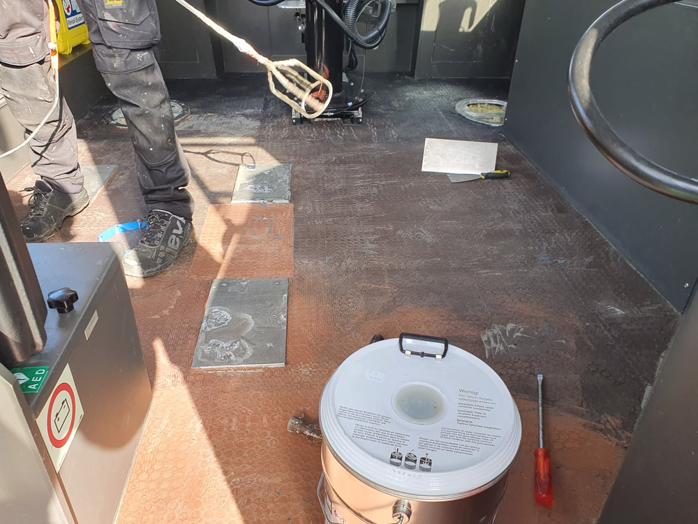 Eine Person steht auf einem staubigen Boden und benutzt ein Rührwerkzeug; auf dem Boden liegen ein großer Metallbehälter und ein Schraubenzieher. Sonnenlicht fällt herein und hebt die Struktur des Bodens sowie die verschiedenen, verstreut liegenden Werkzeuge hervor.