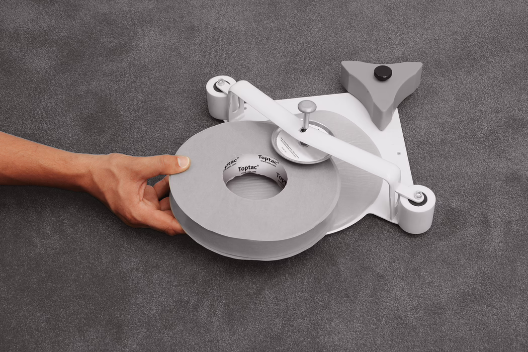 Nahaufnahme einer linken Hand, die eine große graue Klebebandrolle hält, die auf einem flachen Abroller auf einer grau strukturierten Stoffoberfläche liegt. Der Abroller besitzt einen weißen Metallarm mit einer mittigen Spindel und einem runden Schneidrad, zwei kleine weiße Rollen an der Basis sowie einen dreieckigen grauen Stabilisator mit schwarzem Gummifuß; der Pappkern des Bandes zeigt die aufgedruckte Marke 'Toptac'. Weiches, gleichmäßiges Licht betont die neutrale Farbpalette aus Grautönen und Weiß. Die Komposition ist auf die Bandrolle und die Hand unten links zentriert und zeigt Hautfarbe und Adern ohne identifizierende Merkmale.
