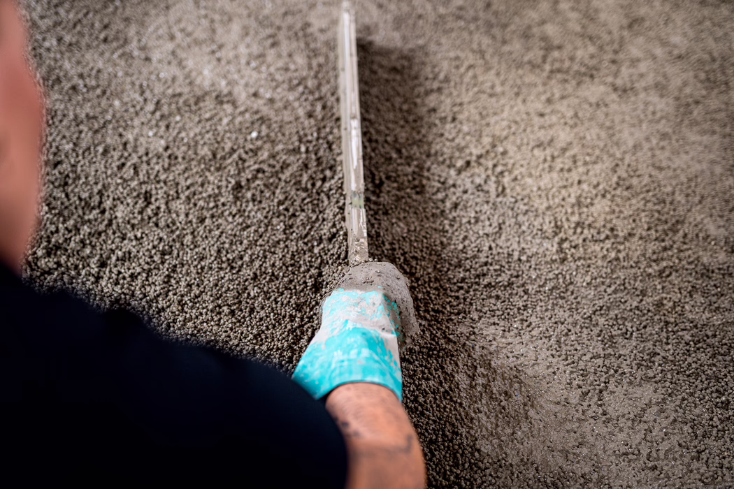 UZIN_Application Image_Screed_Turbolight-System_2023_02 (93) Nahaufnahme von oben: Der Fuß eines Bauarbeiters mit türkisfarbener Schutzschuh-Besohlung führt eine lange, gerade Metalllatte (Abziehlatte) über frisch gegossenen, feuchten Beton oder Ausgleichsmasse, um die Oberfläche zu glätten; die körnige, feuchte Struktur füllt das Bild, die Latte zieht eine glatte Spur durch das Material, geringe Schärfentiefe betont Werkzeug und Oberfläche.