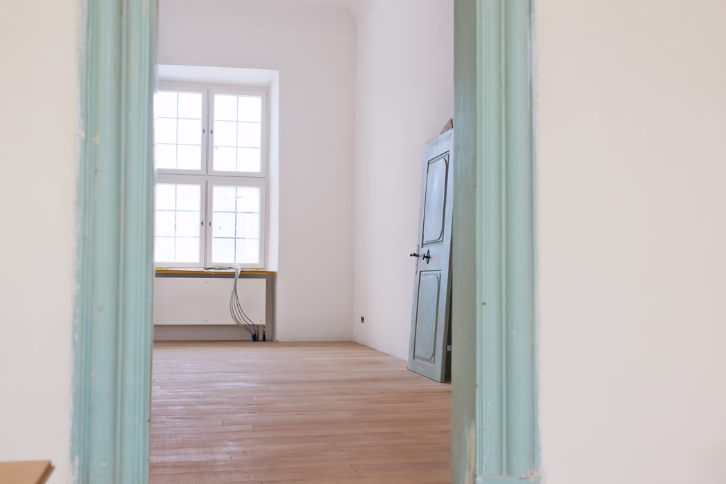 Blick durch einen türkisfarben gestrichenen Türrahmen in einen leeren, sonnendurchfluteten Raum. Ein großes vierfach unterteiltes Fenster in der Mitte der Rückwand flutet den Raum mit sanftem Tageslicht. Der Raum hat frisch geschliffene, helle Holzdielen, weiße Putzwände und eine leicht verwitterte, blau-graue Holztür, die an der rechten Wand angelehnt ist. Nahe dem Fenster liegen niedrige Heizungsrohre und ein Bündel freiliegender Elektrokabel auf der Fensterbank und führen zum Boden, was auf laufende Renovierungsarbeiten hindeutet. Die Gesamtstimmung ist ruhig und minimalistisch, mit gedämpften Pastelltönen und klaren architektonischen Linien.
