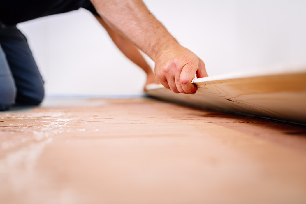 UZIN_Application_Image_MK_250_11_2021-11 Detailaufnahme einer Person, die Boden verlegt: Die Hand eines knieenden Handwerkers fasst die Kante einer Laminat- oder Holzplanke, während sie auf den vorbereiteten Unterboden gelegt wird. Die geringe Schärfentiefe betont die strukturierte Plankenkante und die Hand, im Hintergrund ist eine weiße Wand und die kniende Haltung erkennbar. Die Oberfläche darunter wirkt wie eine Unterlage oder Klebeschicht mit leicht rosa Tönung; die Szene vermittelt sorgfältige manuelle Arbeit bei einer Renovierung.
