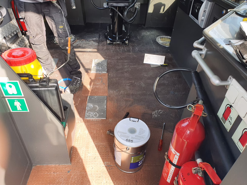 Nahaufnahme des Fahrzeuginnenraums mit einem Feuerlöscher, einer Farbdose, Werkzeugen und Notfallschildern. Eine Person in Arbeitskleidung steht neben der Ausrüstung auf einem braunen Boden. Sonnenlicht wirft Schatten über die Szene.