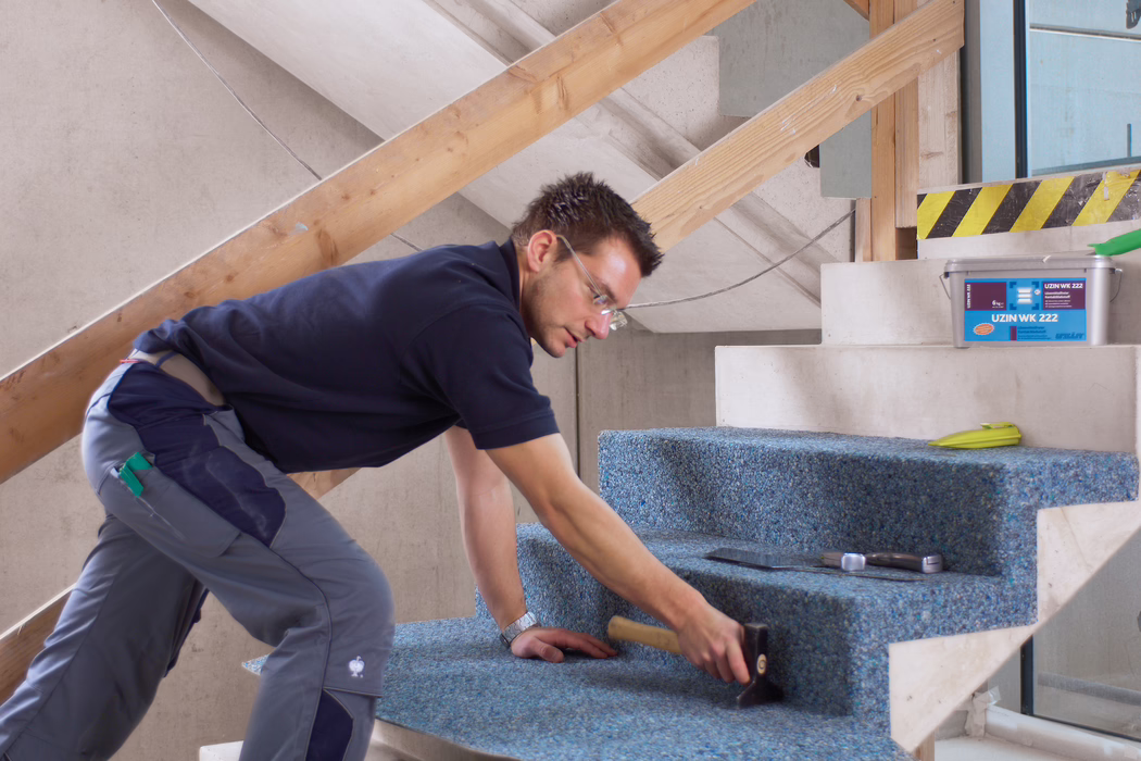 UZIN_Application_Image_WK 222_VA_4_ 2018-03 Ein Handwerker verlegt einen blauen, recycelten Teppichbelag auf eine unvollendete Betontreppe in einem renovierungsbedürftigen Innenraum. Er kniet und beugt sich nach vorn, trägt eine Schutzbrille, ein dunkelblaues Polohemd und graue Arbeitshosen und verwendet einen Gummihammer, um den Teppich fest auf den Stufen anzubringen; in der Nähe liegen eine Glättkelle, ein Maßband und ein Plastikeimer mit der Aufschrift ‚UZIN WK 222‘. Die Szene ist hell mit natürlichem Licht ausgeleuchtet, provisorische Holzbretter als Geländerstützen sind sichtbar, der Schwerpunkt liegt auf der sorgfältigen handwerklichen Arbeit und der Textur des Teppichs auf den Stufentritten.